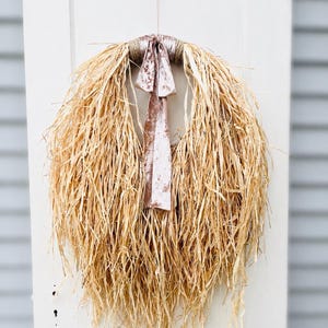 May include: A circular wreath constructed from natural tan raffia fibers, featuring a long, flowing, and textured appearance. At the top center, a dusty rose velvet ribbon is tied in a bow, with two long tails hanging down over the raffia strands.