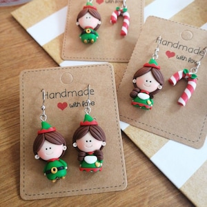 May include: Two pairs of handmade Christmas elf earrings. Each pair features a small elf figurine and a candy cane. The earrings are on brown card stock with the text "Handmade with love".