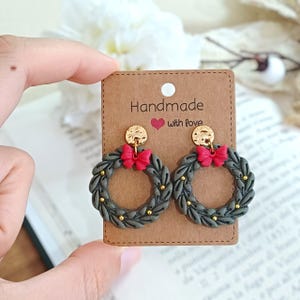 Könnte beinhalten: Handgemachte Kranzohrringe mit roter Schleife und goldenen Akzenten. Die Ohrringe sind grau und goldfarben, mit einem Kranzdesign. Auf der braunen Karte steht "Handmade with love".