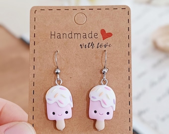 May include: A pair of dangle earrings featuring pink ice cream popsicles with white frosting and colorful sprinkles. The earrings are attached to silver-colored hooks and are displayed on a brown card that reads "Handmade with love."