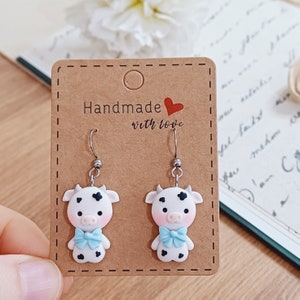 Puede incluir: Un par de pendientes con forma de vaca con manchas blancas y negras, lazos azules y ganchos plateados. Los pendientes se muestran en una tarjeta marrón con el texto "Handmade with love".