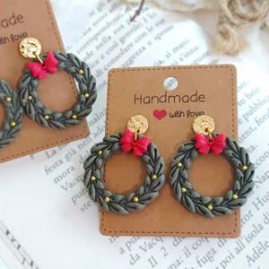 May include: Gray wreath earrings with red bows and gold accents. The earrings are handmade and feature a circular design with small gold beads. The earrings are displayed on a brown card that says "Handmade with love."