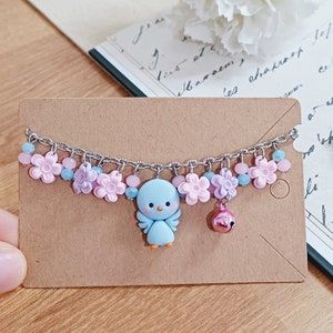 Puede incluir: Un brazalete de cadena plateada con abalorios de flores rosas y moradas, un abalorio de pájaro azul y un abalorio de campana rosa.