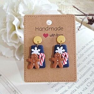May include: Handmade Christmas earrings with a gingerbread man, candy cane, and snowflake design. The earrings have a navy blue background with gold accents, displayed on a brown card.