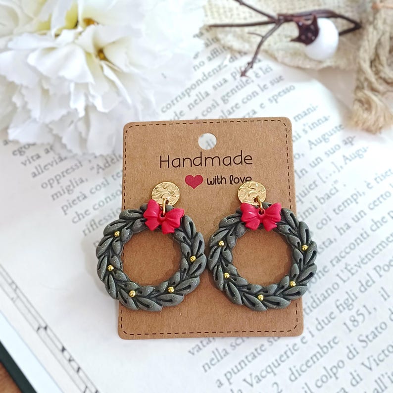 May include: Handmade earrings featuring a gray wreath design with gold accents and a red bow. The earrings are attached to a brown card with the text "Handmade with love."