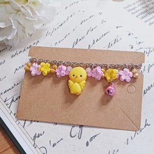 Puede incluir: Un brazalete de cadena plateada con dijes de flores rosas y amarillas, un dije de pollito amarillo y un dije de campana rosa.