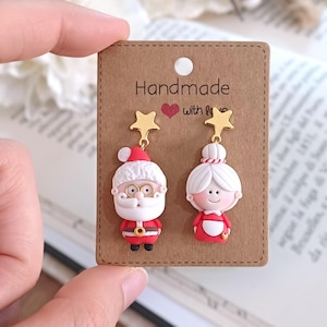 Puede incluir: Pendientes navideños con diseños de Papá Noel y Mamá Noel. El pendiente de Papá Noel tiene un gorro rojo, barba blanca y gafas doradas. El pendiente de Mamá Noel tiene un moño blanco y un vestido rojo. Ambos pendientes tienen detalles de estrella dorada y están en una tarjeta marrón con "Handmade with love."