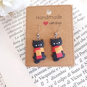 Puede incluir: Pendientes de gato negro hechos a mano con bufandas naranjas y rojas. Los pendientes están en una tarjeta marrón con el texto "Handmade with love" y un corazón. El fondo es un libro.