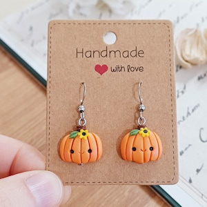 Może przedstawiać: Para handmade'owych kolczyków w kształcie dyni z uśmiechniętą buźką, małym zielonym listkiem i żółtym kwiatkiem na każdej dyni. Kolczyki znajdują się na brązowej kartce z napisem "Handmade with love".
