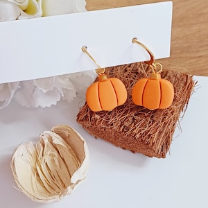 Peut inclure: Une paire de boucles d'oreilles créoles dorées avec des breloques en forme de citrouille orange. Les boucles d'oreilles sont présentées sur une surface brune texturée.