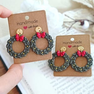 May include: Handmade earrings featuring a wreath design with a red bow, gold accents, and a gold disc. The earrings are displayed on a brown card with the text "Handmade with love".