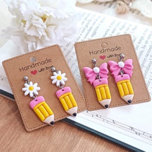 May include: Handmade earrings with a pencil and daisy or bow design. The earrings are yellow, pink, and white, with the text "Handmade with love" on the backing card.