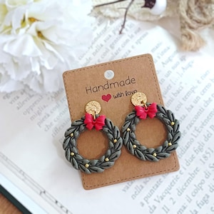 May include: Handmade earrings featuring gray wreath designs with red bows and gold accents. The earrings are displayed on a brown card with the text "Handmade with love".