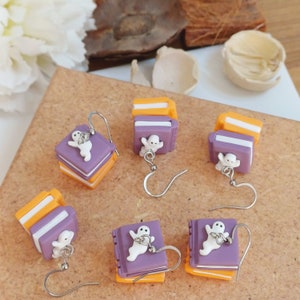 May include: A set of six Halloween-themed earrings featuring a stack of books with a ghost on top. The books are purple and orange, and the ghosts are white. The earrings are on a brown surface.