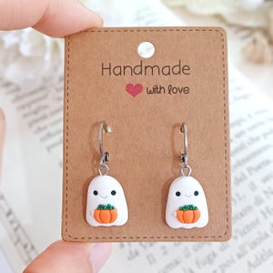 Könnte beinhalten: Ohrringe mit Halloween-Motiv. Jeder Ohrring zeigt einen weißen Geist, der einen orangefarbenen Kürbis hält und an silberfarbenen Creolen befestigt ist. Die Präsentationskarte ist braun und trägt den Text "Handmade with love".