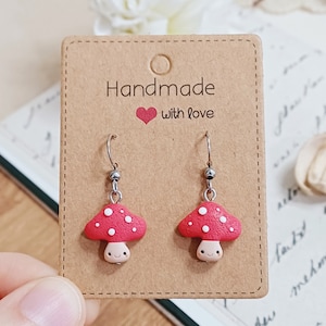 Può includere: Orecchini pendenti a forma di fungo rosso e bianco fatti a mano con un viso sorridente su una carta marrone con il testo "Handmade with love".
