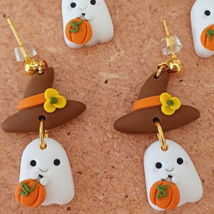 May include: A pair of Halloween-themed earrings featuring a brown witch hat with an orange band and yellow flower accents, each hat is attached to a white ghost holding a small orange pumpkin.