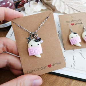 Puede incluir: Un conjunto de collar y pendientes hechos a mano con un fantasma blanco con una pancarta rosa "BOO". El collar tiene una cadena plateada y un pequeño amuleto de araña negra. Los pendientes son ganchos plateados con un amuleto de fantasma blanco.