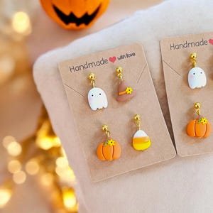Può includere: Orecchini di Halloween fatti a mano con una varietà di design, tra cui un fantasma, un cappello da strega, una zucca e un mais dolce. Gli orecchini sono esposti su una carta marrone con il testo "Handmade with love".