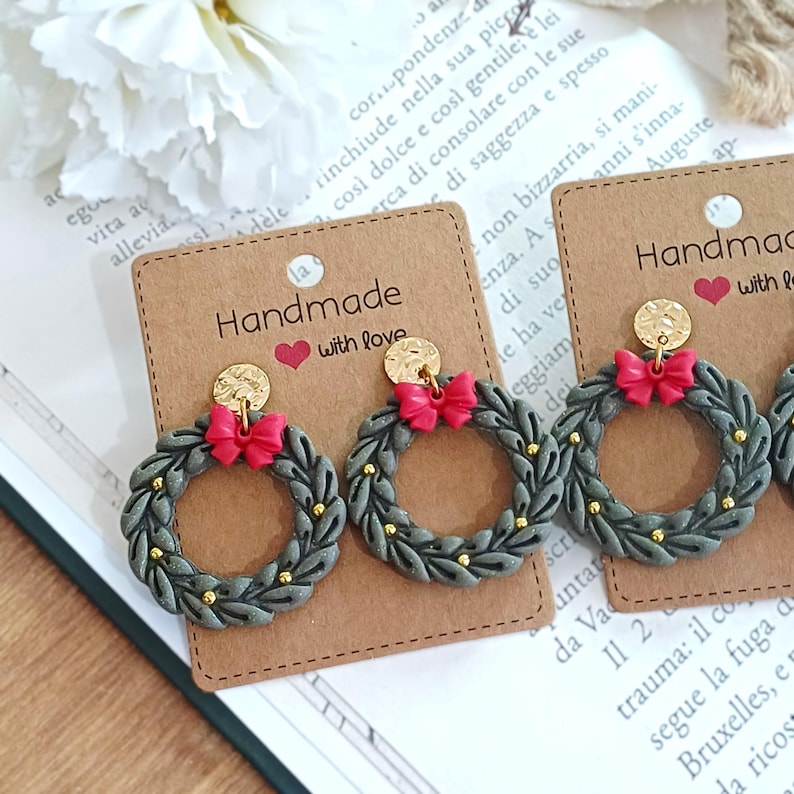 May include: Handmade earrings featuring a gray wreath design with red bows and gold accents. The earrings are attached to a gold-colored disc and are displayed on a brown card with the text "Handmade with love."