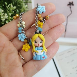 May include: A silver chain bracelet with blue, yellow, and white flower charms. The charm at the end of the chain is a small figurine of Alice in Wonderland wearing a blue dress with a white apron and a red heart.