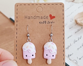May include: A pair of dangle earrings featuring pink popsicle charms with white frosting and colorful sprinkles. The charms have a happy face and a stick. The earrings are on a brown card with the text "Handmade with love".