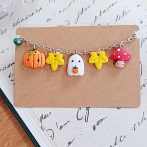 Könnte beinhalten: Ein silbernes Kettenarmband mit herbstlichen Anhängern. Zu den Anhängern gehören ein orangefarbener Kürbis, gelbe Ahornblätter, ein weißes Gespenst mit einem Kürbis und ein roter Pilz mit weißen Punkten. Das Armband wird auf einer braunen Karte präsentiert.
