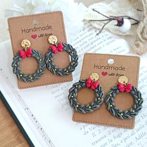 May include: Handmade wreath earrings with a gray base, gold accents, and a red bow. The earrings are displayed on a brown card with the text "Handmade with love". The earrings are a festive accessory.