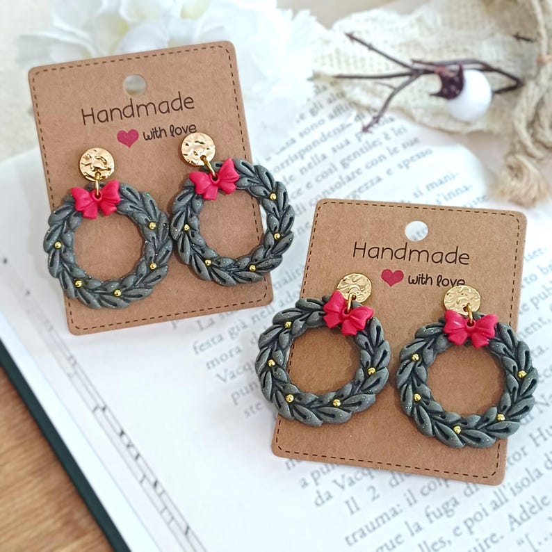 May include: Handmade wreath earrings with a gray base, gold accents, and a red bow. The earrings are displayed on a brown card with the text "Handmade with love". The earrings are a festive accessory.