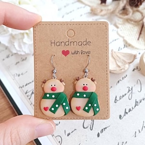 Peut inclure: Boucles d'oreilles de renne de Noël faites à la main avec des écharpes vertes et blanches à pois sur une carte marron avec le texte "Handmade with love".
