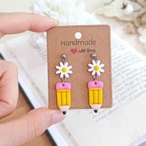 Könnte beinhalten: Ein Paar handgefertigte Ohrringe mit einem gelben Bleistift-Design mit rosa Radiergummi und schwarzer Spitze, gekrönt von einer weißen Gänseblümchen mit gelber Mitte. Die Ohrringe werden auf einer braunen Karte mit der Aufschrift "Handmade with love" präsentiert.