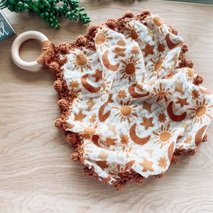 May include: A white muslin baby blanket with a brown crocheted border and pom-poms. The blanket features a repeating pattern of suns and moons in a burnt orange color. A wooden teething ring is attached to the blanket.