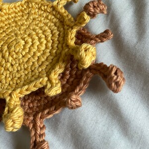 May include: A crocheted sun coaster with a yellow center and brown rays. The coaster is made of yarn and has a textured surface.