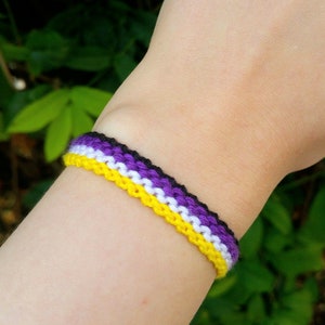Pode incluir: Um bracelete de amizade roxo, branco e amarelo no pulso de uma pessoa.