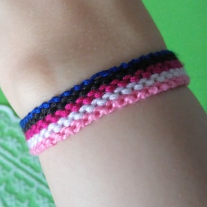 Puede incluir: Un brazalete de amistad multicolor con hilos azules, negros, rosas y blancos tejidos juntos.