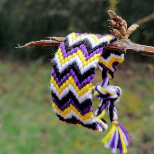 Pode incluir: Um bracelete de amizade roxo, amarelo e preto com um padrão chevron. O bracelete está amarrado a um galho.