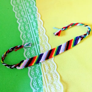 Op de afbeelding: Een kleurrijk vriendschapsarmband met een regenboogpatroon. Het armband is gemaakt van geweven touw en heeft een verscheidenheid aan kleuren, waaronder rood, oranje, geel, groen, blauw, indigo en violet.
