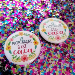 Peut inclure: Deux boutons ronds blancs avec une bordure florale. Le texte sur les boutons est "Le Patriarcat C'est Caca".