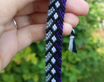 May include: A friendship bracelet with a grey, white, black, and purple color pattern. The bracelet is made with a knotting technique.