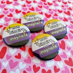 Può includere: Quattro spille rotonde con il testo "non binaire pas imaginaire" in francese. Le spille sono gialle, viola e nere, che rappresentano la bandiera del pride non binario.