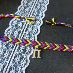Op de afbeelding: Een vriendschapsarmband met een zwart, geel, roze en blauw chevron-patroon. De armband heeft een zilveren bedel met de Griekse letter Pi.