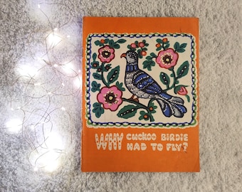 Könnte beinhalten: Ein orangefarbener Buchumschlag mit einer Illustration im Folk-Art-Stil eines blau-grauen Vogels auf einem Ast mit rosa Blumen. Der Text "WHY cuckoo BIRDIE HAD TO FLY?" ist in Weiß gedruckt.