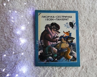 May include: A rectangular children's book with a blue border featuring an illustration of a wolf and a fox holding a fish in a snowy landscape. The text "ЛИСИЧКА-СЕСТРИЧКА І ВОВК-ПАНІБРАТ" is printed in black at the top of the cover, accompanied by a small bird icon.