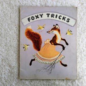 Op de afbeelding: Vintage boekomslag getiteld "Foxy Tricks" met een cartoonvos in een jurk, met een bezem en omringd door gele vogels. De vos heeft een bruin en wit lichaam met een grote, pluizige oranje staart. De achtergrond is lichtpaars.