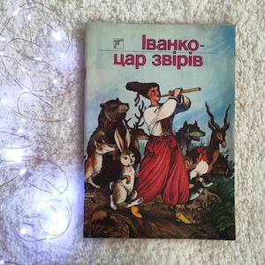 Pode incluir: Um livro ilustrado vintage intitulado Ivanko-tsar zviriv com uma figura em trajes tradicionais tocando flauta, rodeada por um urso, um coelho, um lobo e um veado numa floresta, ao lado de um fio de pequenas luzes LED brancas.