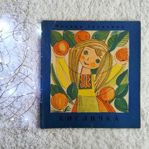 Puede incluir: Un libro infantil vintage con una cubierta azul. La ilustración muestra a una niña sonriente con cabello rubio, sosteniendo naranjas, rodeada de hojas verdes y naranjas. El título del libro y el nombre del autor son visibles.