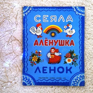 Peut inclure: Un livre pour enfants vintage avec une couverture bleue ornée d'illustrations colorées et de texte cyrillique. La couverture comprend des images d'un coq, d'un arc-en-ciel, d'une oie et d'une femme en robe rouge. Le titre du livre est en grandes lettres blanches.