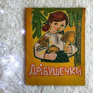 Puede incluir: Libro infantil vintage con una cubierta amarilla. La ilustración muestra a una niña con cabello rojo, vestida con una camisa bordada tradicional y una falda verde, sosteniendo champiñones. El título "ДРІБУШЕЧКИ" está escrito en letras rojas.
