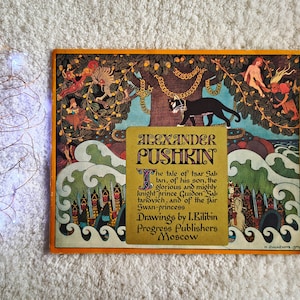Op de afbeelding: Een vintage boekomslag met de titel "Alexander Pushkin" en illustraties. De omslag heeft een oranje rand en toont een zwarte kat, figuren en een boom met gouden kettingen. De tekst bevat de titel en de zin "Het verhaal van tsaar Saltan".