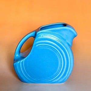 May include: A vibrant turquoise ceramic pitcher with a rounded body and a handle. The pitcher features concentric circle detailing and a spout for pouring. The background is a gradient of orange and tan.